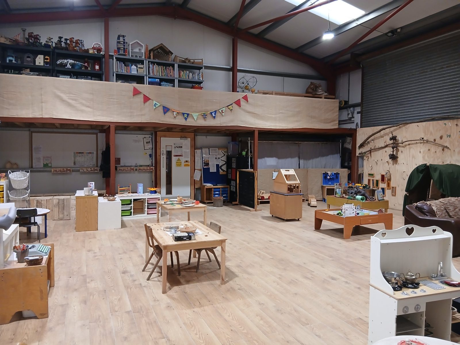 Inside the main hall at Evergreen Forest Nursery, with Evergreen bunting, play tables, shelves of books and toys, and a wooden feature wall